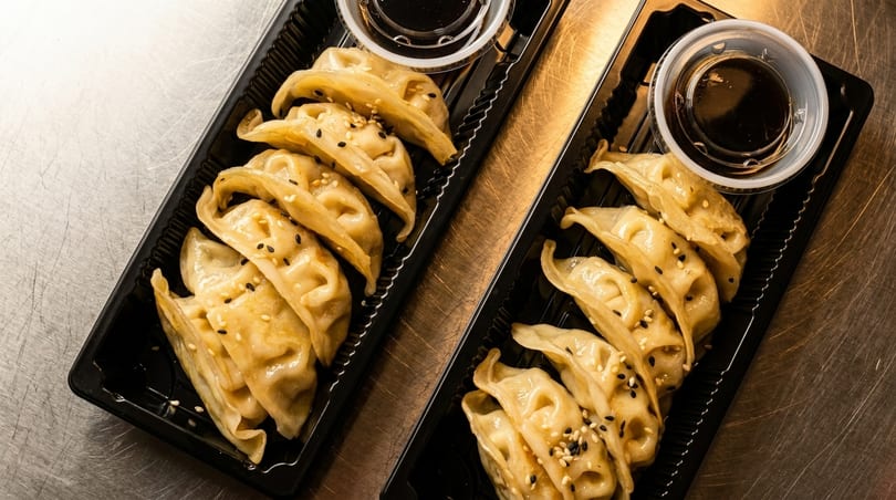 Two black takeout containers filled with golden-brown dumplings topped with sesame seeds and dipping sauce.